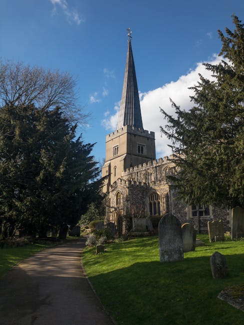 A tall church steeple with a pointed, slate-grey roof rises above a historic stone church building, surrounded by a graveyard with weathered headstones. The church has detailed stonework and arched wi